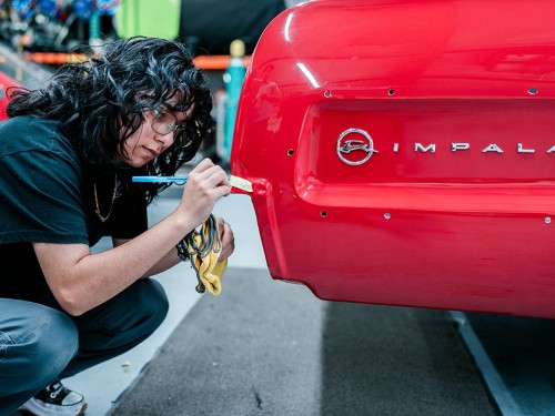 A young person wearing jeans and short sleeve shirt crouches next to end of red Impala.